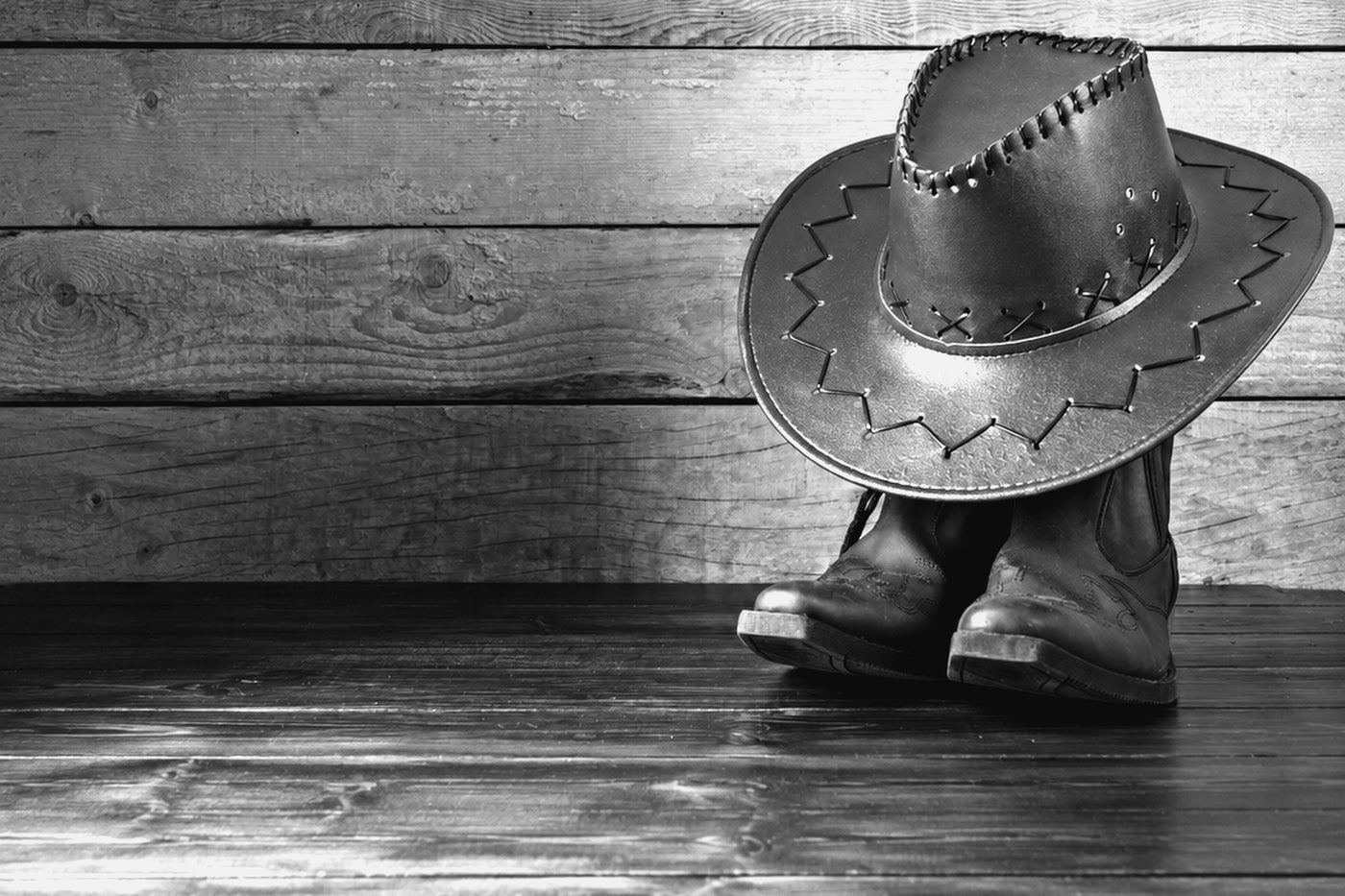 A leather cowboy hat resting on a panel of weathered wood, photographed in black and white.