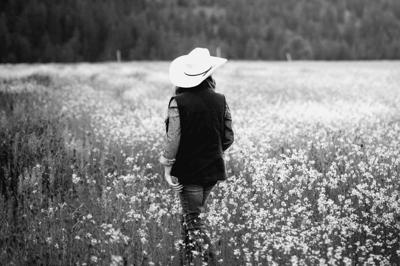 A woman seen from behind in a cowboy hat, standing in a meadow, photographed in black and white.